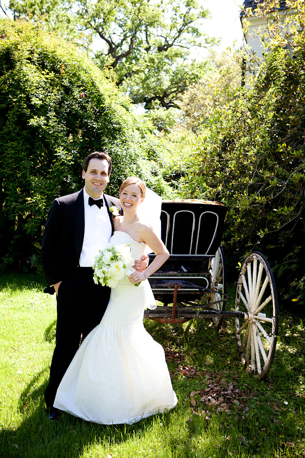 the happy couple standing in front of a carriage in a lawn - bride is wearing a white mermaid style dress holding a white and green bouquet and groom is wearing a black and white tuxedo - photo by North Carolina based wedding photographers Cunningham Photo Artists